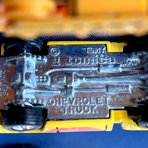 May include: Close-up of a yellow and silver toy Chevrolet truck. The underside of the truck is visible, with the text "CHEVROLET TRUCK", "TOMICA", and "JAPAN" embossed on the metal. The toy has black wheels.