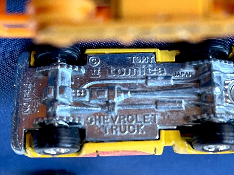 May include: Close-up of a yellow and silver toy Chevrolet truck. The underside of the truck is visible, with the text "CHEVROLET TRUCK", "TOMICA", and "JAPAN" embossed on the metal. The toy has black wheels.