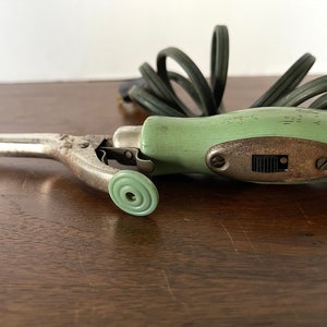 Puede incluir: Un rizador de pelo eléctrico vintage verde y blanco con un mango redondo verde y un cable. El rizador tiene un interruptor de metal en el lateral.
