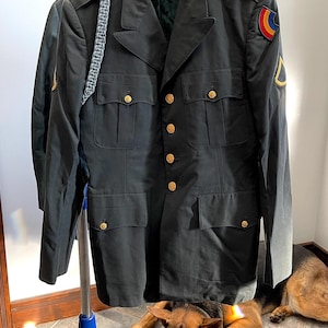 Puede incluir: Una chaqueta de uniforme militar verde oscuro con botones dorados y un parche en la manga. La chaqueta está colgada en un perchero. Un perro pastor alemán está tumbado en el suelo delante de la chaqueta.