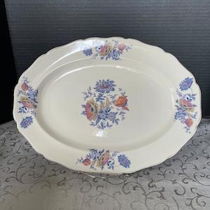 Puede incluir: Un plato ovalado blanco para servir con un borde festoneado y un diseño floral en tonos azules, amarillos y naranjas. El diseño floral presenta flores y hojas estilizadas.