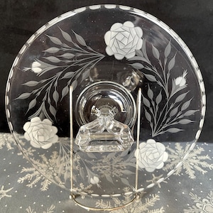 May include: A clear glass pedestal cake plate with an etched design of roses and leaves. The plate has a scalloped edge and a clear glass pedestal.