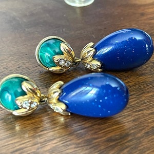 Puede incluir: Un par de pendientes colgantes con piedras azules y verdes. Las piedras azules tienen forma de lágrima y las piedras verdes son redondas. Los pendientes son de tono dorado y tienen un diseño floral.