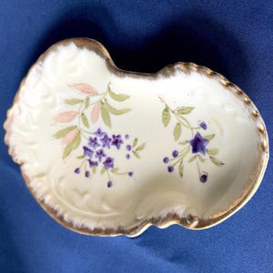 Könnte beinhalten: Eine kleine, cremefarbene Porzellanschale mit gewelltem Rand und Goldrand. Die Schale ist mit handgemalten Blumendesigns in Lila, Rosa und Grün verziert. Die Schale wird wahrscheinlich zum Servieren kleiner Portionen oder als Dekorationsartikel verwendet.