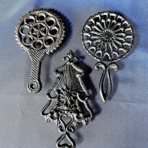 Puede incluir: Tres piezas decorativas de metal plateado. Una es redonda con un diseño de estrella, otra tiene forma de espejo de mano y la tercera es una forma de árbol estilizada con detalles de corazón. Las piezas están dispuestas sobre un fondo azul.