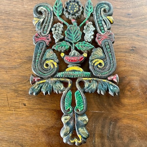 May include: A cast iron trivet with a floral design painted in green, yellow, red, and white. The trivet is shaped like a stylized flower with a stem.