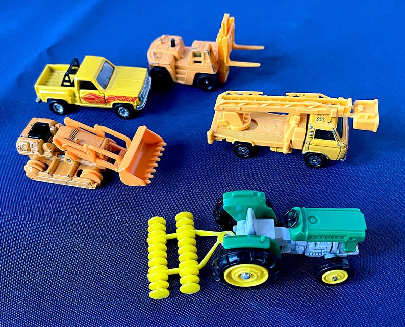 May include: A collection of toy construction vehicles, including a yellow pickup truck, a forklift, a bulldozer, a crane truck, and a green tractor with yellow wheels and a yellow implement. The toys are set against a blue background.