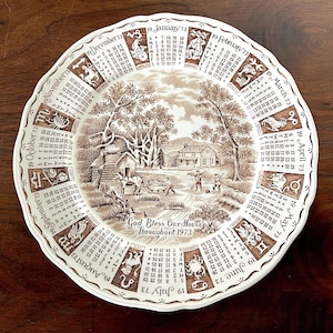 Könnte beinhalten: Ein brauner und weißer Keramikteller mit einem Kalender für jeden Monat des Jahres 1973. Der Teller zeigt eine ländliche Szene mit einem Bauernhaus, Bäumen und Personen. Der Text "God Bless Our House Throughout 1973" ist auf dem Teller aufgedruckt.