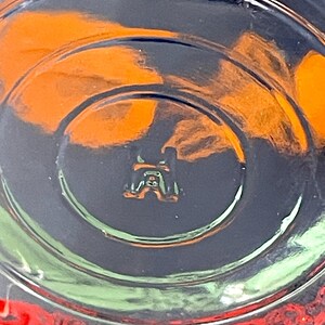May include: Close-up of the bottom of a clear glass with a circular design. The glass has a small, embossed emblem at the center. Orange and red colors are visible through the glass, with a dark background.