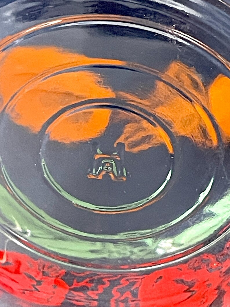 May include: Close-up of the bottom of a clear glass with a circular design. The glass has a small, embossed emblem at the center. Orange and red colors are visible through the glass, with a dark background.