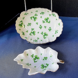 Pode incluir: Dois pratos de cerâmica branca com desenhos de trevos verdes. Um é oval com borda ondulada, e o outro em forma de folha. Ambos têm acabamento dourado. Os pratos estão sobre uma superfície azul.