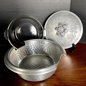 以下が含まれることがあります： ハンドル付きの透明なガラス製ボウル、ハンマー加工された金属製のボウル、花とブドウのデザインのシルバーの蓋。 蓋は黒いスタンドで支えられています。 アイテムは木製の表面にあります。