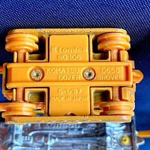 May include: Close-up of the underside of an orange Komatsu Dozer toy. The toy has the text "tomica NO.106" and "KOMATSU DOZER D658 SHOVEL S-1/87 MADE IN JAPAN" printed on it. The toy is made of metal and plastic.