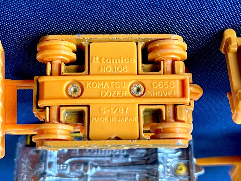 May include: Close-up of the underside of an orange Komatsu Dozer toy. The toy has the text "tomica NO.106" and "KOMATSU DOZER D658 SHOVEL S-1/87 MADE IN JAPAN" printed on it. The toy is made of metal and plastic.