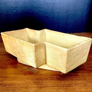 May include: A rectangular ceramic dish with a light cream-coloured glaze and subtle brown speckles throughout. The vessel features a distinct stepped or notched geometric design along its upper rim and sides, creating a unique angular profile.