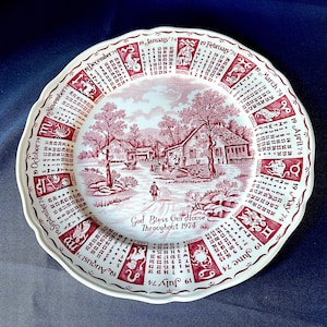 May include: A decorative plate with a red and white design. The plate features a calendar for 1974, zodiac signs, and a rural landscape scene with the text "God Bless Our House Throughout 1974". The plate has a scalloped edge.