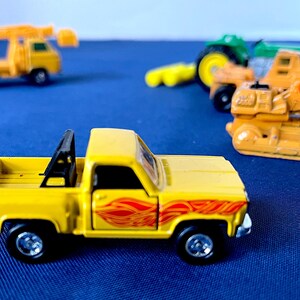 May include: A yellow toy pickup truck with a black roll bar and red flame decals. Other toy construction vehicles are in the background, including a tractor, a forklift, and a crane truck. The toys are on a blue surface.