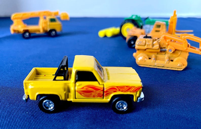 May include: A yellow toy pickup truck with a black roll bar and red flame decals. Other toy construction vehicles are in the background, including a tractor, a forklift, and a crane truck. The toys are on a blue surface.
