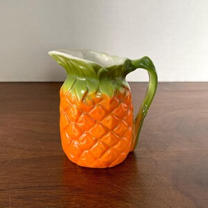 Op de afbeelding: Een keramische kan in de vorm van een ananas met een groene bovenkant en een oranje lichaam. De kan heeft een handvat en een tuit.