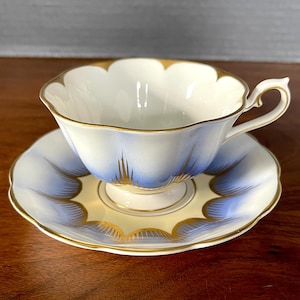 May include: A white teacup and saucer set with a scalloped rim and gold trim. The cup and saucer feature a blue and gold floral design. The teacup has a delicate handle and sits on a matching saucer.