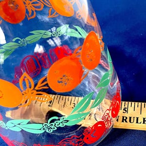 May include: Clear glass jar with orange fruit and green leaf designs. The jar has a wide mouth and a slightly tapered body. A wooden ruler is visible next to the jar. The background is a solid blue.
