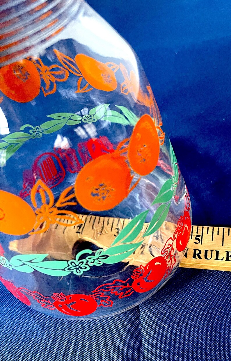 May include: Clear glass jar with orange fruit and green leaf designs. The jar has a wide mouth and a slightly tapered body. A wooden ruler is visible next to the jar. The background is a solid blue.