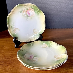 May include: Three round porcelain plates with scalloped edges and a beaded rim. The plates feature a soft cream center, a pale green border, and hand-painted pink and white rose motifs. One plate is displayed upright on a black plastic stand.