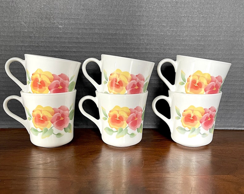 May include: Six white ceramic mugs with a floral design featuring yellow and pink pansies. The mugs are stacked in pairs.