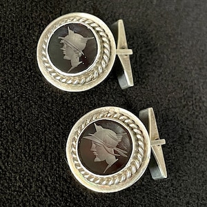 May include: A pair of silver cufflinks with black cameo insets. Each cufflink features a profile of a person wearing a helmet.