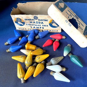 May include: A collection of vintage General Electric Mazda Christmas tree lamps in various colors, including blue, red, yellow, white, and green. The lamps are scattered on a blue surface near their original cardboard packaging.