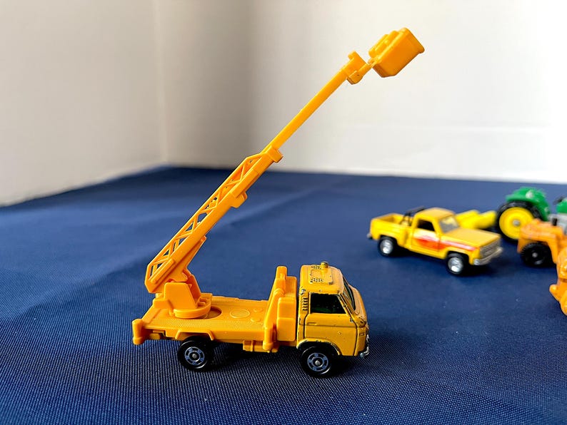 May include: A yellow toy truck with an extending crane arm. The crane arm is raised, and the truck has black wheels. Other toy vehicles are visible in the background.