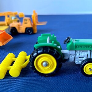 May include: A green and gray toy tractor with black tires and yellow rims, labeled "KUBOTA". A yellow farm implement is attached. Other toy construction vehicles are in the background, including a yellow forklift and a front-end loader.