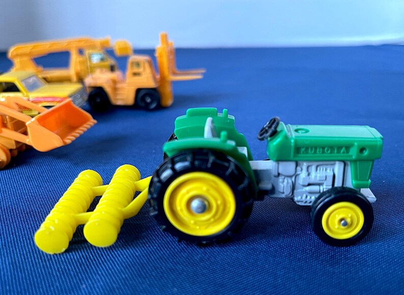 May include: A green and gray toy tractor with black tires and yellow rims, labeled "KUBOTA". A yellow farm implement is attached. Other toy construction vehicles are in the background, including a yellow forklift and a front-end loader.
