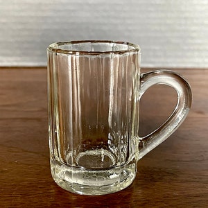 May include: A clear glass mug with a handle and vertical lines. The mug is empty and sitting on a wooden surface.