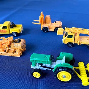 May include: A collection of toy construction vehicles, including a yellow pickup truck, a forklift, a bulldozer, a truck with a ladder, and a green tractor with yellow wheels and a yellow roller attachment. The toys are on a blue surface.