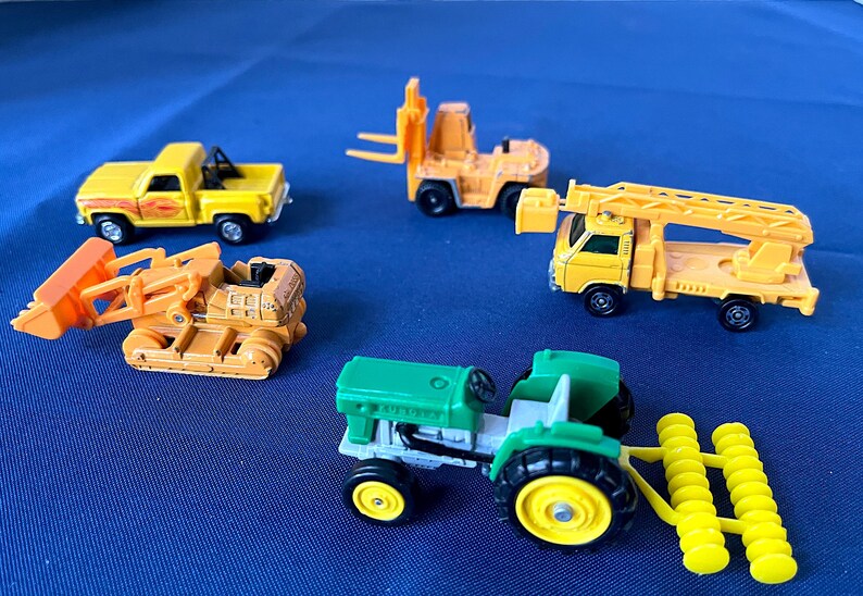 May include: A collection of toy construction vehicles, including a yellow pickup truck, a forklift, a bulldozer, a truck with a ladder, and a green tractor with yellow wheels and a yellow roller attachment. The toys are on a blue surface.
