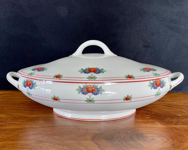 May include: White oval serving dish with a lid, featuring a floral pattern in red, blue, and green. The dish has a red and white trim around the edge.