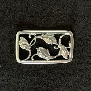 May include: A rectangular silver-toned metal belt buckle with an openwork floral design. The buckle features stylized leaves and stems within a rectangular frame. The design is symmetrical and detailed, with a polished finish.