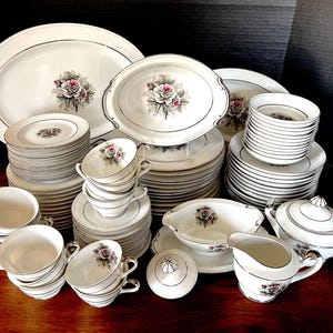 May include: A complete set of white porcelain dinnerware with a delicate floral design featuring pink roses and green leaves. The set includes plates, bowls, cups, saucers, a gravy boat, a creamer, and a sugar bowl.