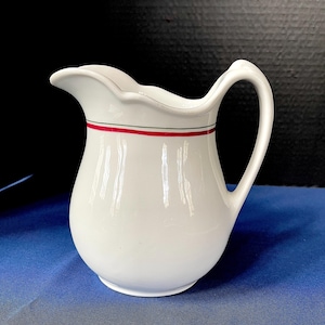 Może przedstawiać: Biały ceramiczny dzbanek z zakrzywionym uchwytem i falistym brzegiem. Dzbanek posiada czerwono-czarny pasek wokół korpusu. Klasyczny design do serwowania napojów.