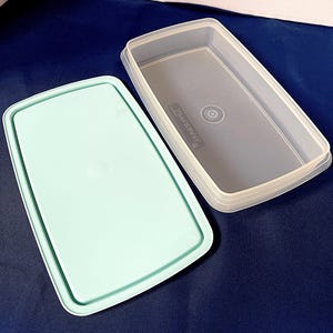 Puede incluir: Un recipiente rectangular de plástico transparente con una tapa azul claro. La tapa está a un lado, revelando el interior del recipiente. El recipiente tiene esquinas redondeadas y una superficie ligeramente texturizada. La tapa y el recipiente son de plástico duradero.