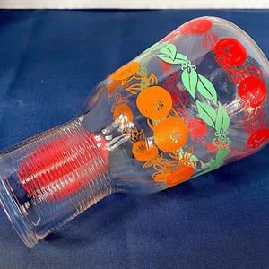 May include: A clear glass pitcher with a wide base and a narrow neck. The pitcher is decorated with orange and red fruit, green leaves, and red floral designs. The base has a red, ribbed design.