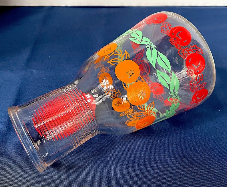 May include: A clear glass pitcher with a wide base and a narrow neck. The pitcher is decorated with orange and red fruit, green leaves, and red floral designs. The base has a red, ribbed design.