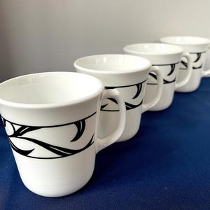 May include: Four white ceramic mugs with a black abstract design. The mugs are arranged in a row, each with a handle and a curved black pattern around the middle. The mugs are on a blue surface.