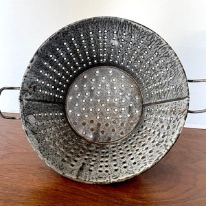 May include: A vintage gray and white enamel colander with a round shape and two handles. The colander has a speckled pattern and a large number of holes.