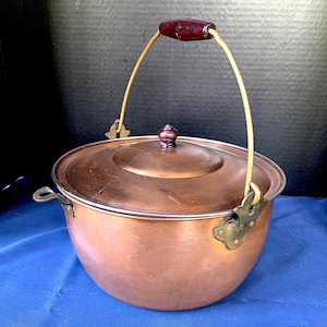 May include: A copper pot with a lid and a handle. The pot has a rounded shape with a shiny finish. The handle is made of a dark wood and is attached to the pot with brass hardware. The pot is sitting on a blue surface.