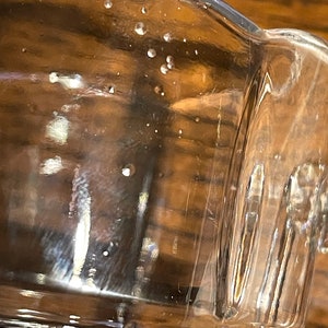 Op de afbeelding: Een helder glazen beker met een getextureerde bodem en een gladde rand. De beker heeft verschillende kleine luchtbellen die in het glas zijn opgesloten.