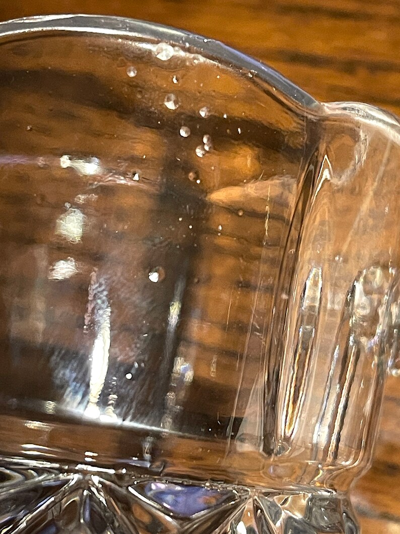Op de afbeelding: Een helder glazen beker met een getextureerde bodem en een gladde rand. De beker heeft verschillende kleine luchtbellen die in het glas zijn opgesloten.