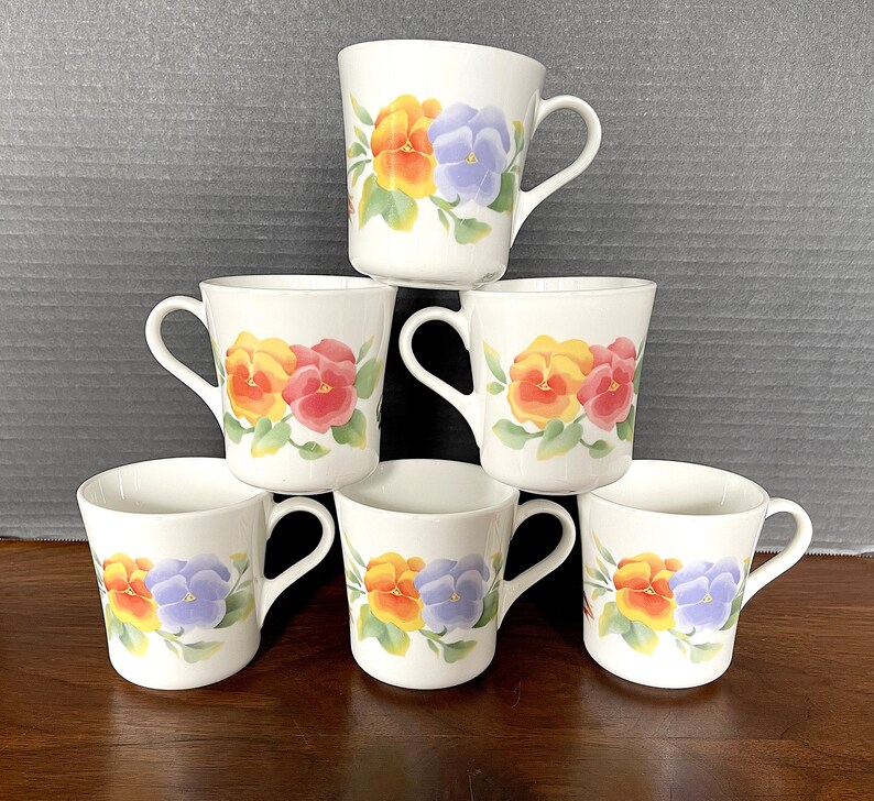 May include: Six white ceramic mugs with handles, each featuring a different floral design with yellow, purple, red, and orange pansies.