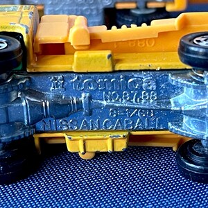 May include: Close-up of a yellow and blue toy truck. The underside of the truck is blue with black wheels. The text "Tomica NO.87,88" and "NISSAN CABALL" are visible.